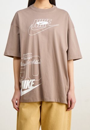 Personne portant un T-shirt Nike marron ample avec des logos et du texte blancs, associé à un pantalon jaune, se tenant devant un fond clair.