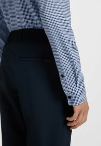 Camicia a maniche lunghe di color azzurro chiaro con un motivo a diamante. Pantaloni scuri con vita elasticizzata. Polsini abbottonati visibili nell'immagine.