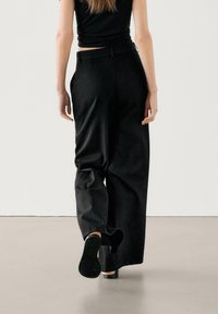 Zwarte high-waisted broek met een losse fit, met zijzakken en een gladde structuur. De persoon draagt zwarte schoenen, waarbij de zool zichtbaar is.