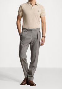 Camisa polo beige con cuello y logo bordado, combinada con pantalones grises con pliegues y zapatos de cuero marrón.