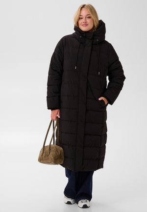 Femme portant un long manteau matelassé noir avec capuche, tenant un sac à main vert olive, debout avec un pantalon bleu et des baskets blanches.