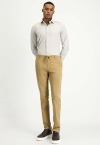 Helles graues Langarmshirt kombiniert mit khakifarbenen Bundhose mit Kordelzug, im entspannten Schnitt. Das Modell trägt dunkle Slipper.