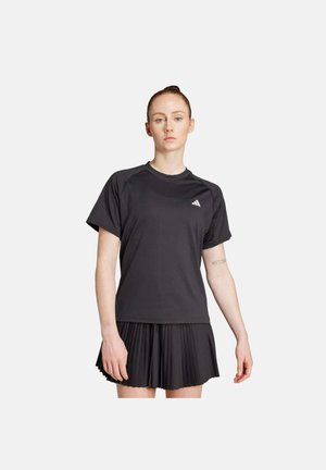 Mujer de pie con expresión neutral, vestida con una camiseta deportiva negra y una falda negra plisada sobre un fondo blanco liso.