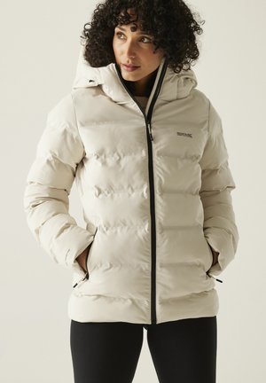 Mujer con cabello rizado que lleva una chaqueta acolchada con capucha de color beige claro, con cremallera negra y las manos en los bolsillos, mirando hacia un lado.