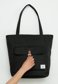 Černá látková taška přes rameno s dvěma dlouhými uchy, předním klopou, hnědým koženým detailem a kovovou přezkou. Logo Herschel viditelné v pravém dolním rohu.