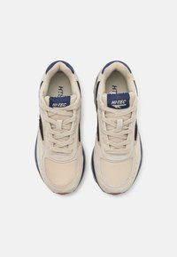 Adidași bej cu un superior din plasă, accente din suede, detalii albastre și o talpă cu amortizare. Sforile și brandingul sunt evidențiate.