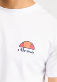 Weißes Baumwoll-T-Shirt mit einem bunten Logo in Orange und Marineblau, das auf der linken Brustseite platziert ist. Mit kurzen Ärmeln.