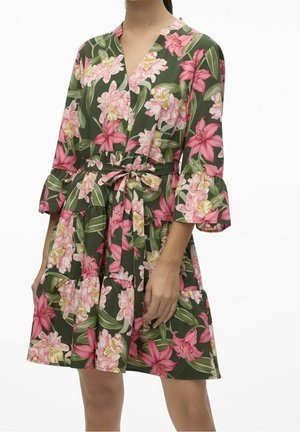 Kobieta w zielonej sukience do kolan w różowo-białe kwiaty, z dekoltem w serek, rękawami do łokcia z falbanami oraz wiązanym paskiem w talii.