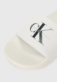 Λευκή σανδάλι με υφασμάτινη λωρίδα που φέρει το μαύρο λογότυπο "Calvin Klein". Η σόλα είναι κατασκευασμένη από υλικό με υφή και λεία επιφάνεια.