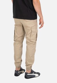 Hombre con pantalones cargo beige con puños elásticos y zapatillas negras Puma, de pie, visto desde atrás sobre un fondo blanco.