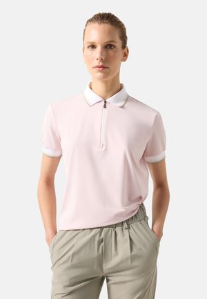 Femme debout, les mains dans les poches, portant un polo à manches courtes rose clair avec un col blanc et un pantalon beige.