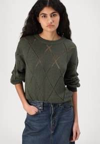 Olivgrüner Strickpullover mit Rautenmuster, hochgerollten Ärmeln und weitem Ausschnitt, kombiniert mit einem blauen Denim-Rock.