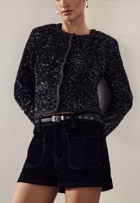 Svart paljettjacka med rund halsringning och kort design, kombinerad med svarta högmidjade manchestershorts med framfickor och bälte.