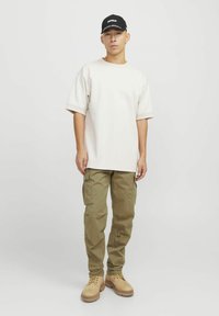 Beige übergroßes T-Shirt mit kurzen Ärmeln, olivgrüne Cargo-Hosen und beige Stiefel. Schwarze Mütze mit weißer Schrift. Einfaches, lässiges Outfit.