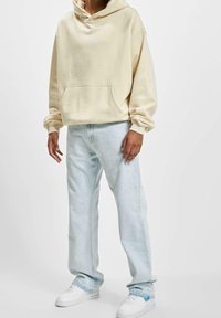 Sudadera beige claro con un gran bolsillo frontal y un corte relajado, combinada con jeans azul claro y zapatillas blancas. Tejido suave, diseño casual.