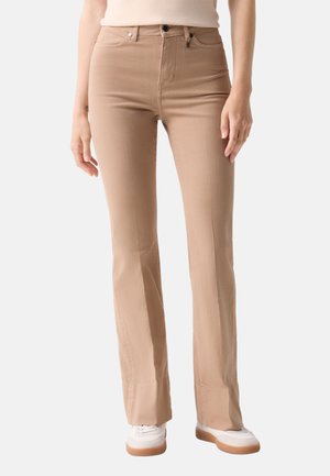Beige bukser med lige ben båret af en person iført hvide sneakers og en lys top, stående mod en ensfarvet baggrund.
