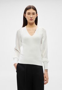 Γυναίκα με μακριά σκούρα μαλλιά που φοράει λευκό πουλόβερ με V-λαιμόκοψη και διαφανείς φουσκωτές μανίκες, καθώς και μαύρο παντελόνι, στέκεται μπροστά σε απλό ανοιχτό φόντο.