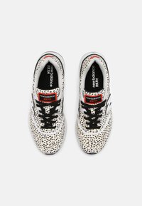 Baskets blanches avec des éclats noirs, dotées de lacets noirs, d'une étiquette rouge et blanche, et d'une semelle rembourrée. Marque : New Balance, style 997H.