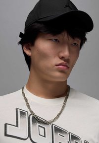 Gorra negra con un logo, collar de cadena plateada y camiseta blanca con gráfico. Enfoque en el rostro con una expresión neutral y cabello texturizado.