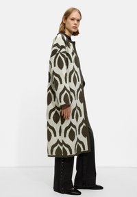 Long cardigan with a green and white leaf pattern, crafted from a textured knit. Features dropped shoulders and a straight fit. Black trousers complete the look.