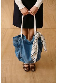 Inimene hoiab käes suurt sinist teksakoti valgete nöörkäepidemete ja valge mustriga salliga, kandes musta seelikut ja leopardimustrilisi madalaid kingi.