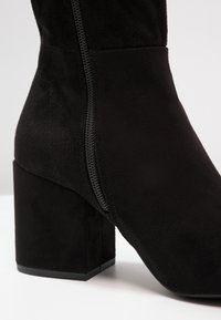 Černé semišové kotníkové boty s blokovým podpatkem, bočním zipem a hladkým texturem. Design zdůrazňuje jednoduchý, moderní tvar.