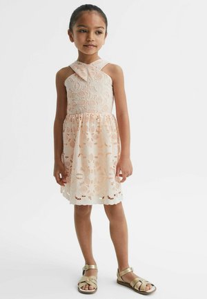 Nuori tyttö hihattomassa vaaleanpunaisessa pitsimekossa, jossa on kukkakuosi, ja kultaiset sandaalit seisoo yksivärisen taustan edessä.