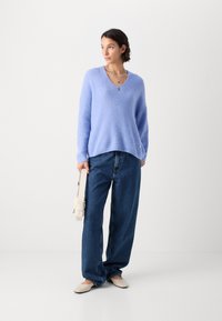 Lichtblauwe gebreide trui met V-hals, relaxte pasvorm, gecombineerd met donkerblauwe wijdgezomen jeans. Geaccentueerd met gelaagde zilveren halskettingen.