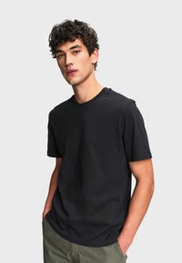 Jeune homme aux cheveux bouclés portant un t-shirt noir à manches courtes et un pantalon vert, debout les mains dans les poches devant un fond gris.