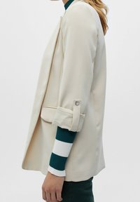 Blazer beige aux manches retroussées, avec une texture lisse et une coupe ajust ée. Porté par-dessus un pull côtelé rayé vert et blanc.