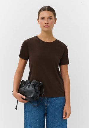 Femme portant une chemise à manches courtes marron foncé et un jean bleu, tenant un sac à cordon en cuir noir sur un fond clair uni.