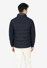 Chaqueta acolchada azul navy con costuras horizontales, cuello alto y detalle del logo en la parte trasera del cuello. El material parece suave e isolado.