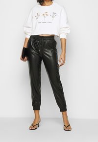 Vit kortärmad sweatshirt med blommönster och text, parat med svarta joggers i konstläder med elastisk midja och manschetter.