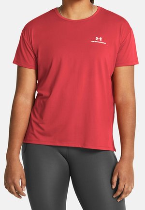 Personne portant un t-shirt rouge à manches courtes Under Armour et des leggings gris foncé, debout avec les bras détendus le long du corps.