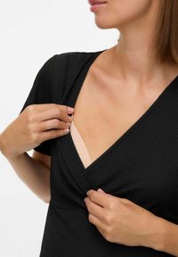 Haut noir texturé avec un décolleté en V. Le tissu présente des motifs subtils. Un sous-vêtement nude est partiellement visible sous le col.