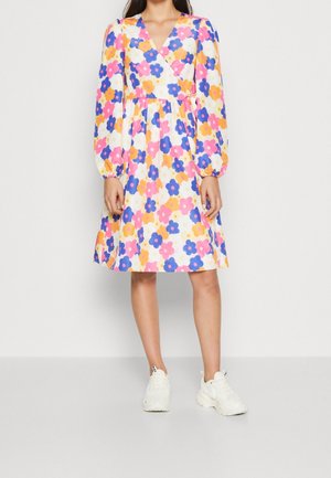 Kvinde iført en knælængde kjole med lange ærmer og flerfarvet blomstermønster, parret med hvide sneakers, stående på en ensfarvet baggrund.