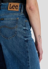 Μπλε τζιν από denim με πίσω τσέπη και καφέ ετικέτα Lee, που φοριέται από άτομο με μικρό λουλουδένιο τατουάζ στον πήχη.