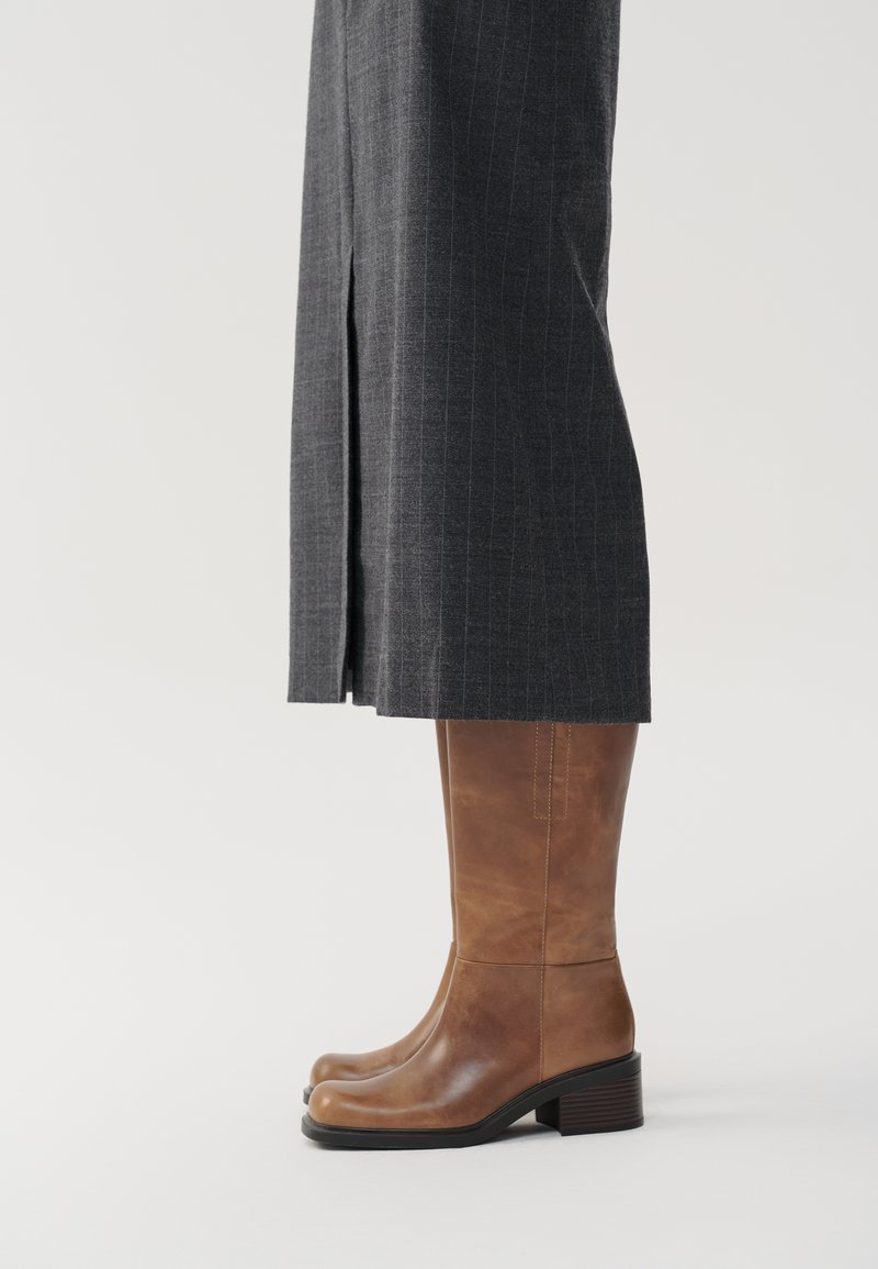 Stivali alti in pelle marrone con punta arrotondata e tacco spesso, abbinati a una gonna grigia a righe verticali con spacco laterale.