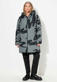 Parka impermeable gris con patrones abstractos negros, que cuenta con capucha, cremallera frontal, dos bolsillos laterales y un diseño hasta la rodilla.