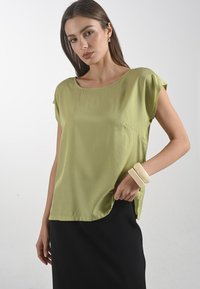 Světle zelený top s krátkým rukávem a kulatým výstřihem, se bočními kapsami, spárovaný s černou sukní a béžovými elastickými náramky na jedné ruce.