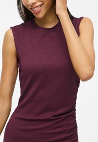 Burgundy ärmlös topp med en texturerad yta. Har en rund halsringning och en figurnära silhuett med rynkade detaljer vid midjan.