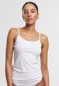 White cotton tank top with thin shoulder straps, fitted design, smooth texture, and no visible patterns or embellishments.