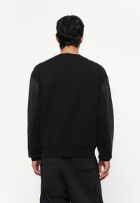 Hanorac negru cu guler rotund și mâneci lungi. Prezintă un material texturat de culoare neagră pe mâneci. Design simplu, croială lejeră.