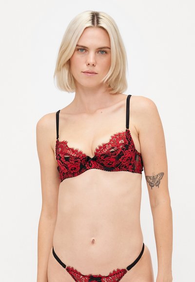 Soutien-gorge en dentelle rouge avec des motifs floraux, des bretelles noires et un petit nœud noir au centre. Texture douce avec des détails semi-transparents.