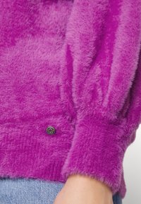 Luddig lila tröja med en texturerad yta, som har puffärmar och en svart knappdetalj vid manschetten. Mjukt stickat material.