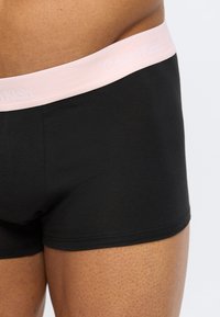 Κοντινό πλάνο ενός ατόμου που φοράει μαύρα αντρικά μποξεράκια Calvin Klein με ροζ α νοιχτή ζώνη μέσης σε μονόχρωμο φόντο.