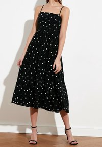 Kobieta w czarnej, bezrękawnikowej sukience midi w białe gwiazdy i czarnych szpilkach na kostkę stoi na drewnianej podłodze.