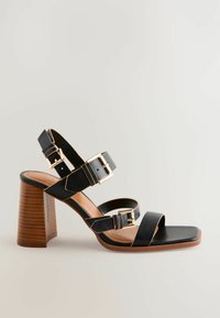 Sandali con tacco neri e punta quadrata, caratterizzati da più cinturini e fibbie in tonalità oro. Il tacco ha un design in legno texturizzato.