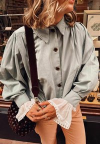 Chemise oversize bleu clair avec un bouton devant, des manches plissées et des poignets en dentelle blanche. Sac épaule en suede marron foncé avec des accents métalliques.