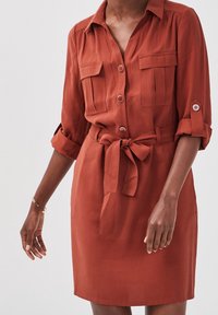 Robe couleur rouille avec manches retroussées, fermeture à boutons sur le devant, poches poitrine et ceinture nouée à la taille, portée par une personne à la peau foncée sur un fond uni.
